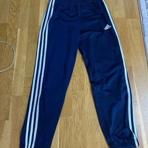 Blå Adidas träningsbyxor  - Snygga marinblå träningsbyxor från Adidas med klassiska vita ränder längs sidorna och logga på låret. Byxorna har elastisk midja med blå snörning och är gjorda i mjuk polyester som ger skön passform. Perfekta för sport eller chill. Inga fickor. Använts 1 gång 