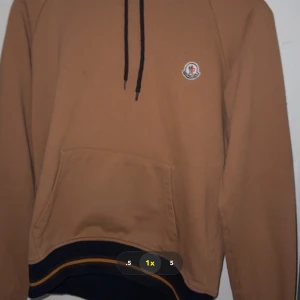 Beige hoodie från Moncler med huva L-M - Säljer en beige hoodie från Moncler i storlek L. Tröjan har huva med snörning, en stor magficka och ribbade muddar med mörkblå och gula ränder. Moncler-logga på bröstet. Perfekt för dig som gillar stilrena och exklusiva streetwear-plagg.
