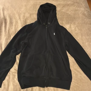 Svart hoodie från Polo Ralph Lauren - Svart hoodie från Polo Ralph Lauren med klassisk broderad logga på bröstet. Tröjan har dragkedja framtill, huva med snörning och stora fickor. Perfekt för en chill och stilren look.