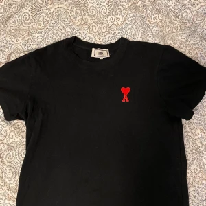 Svart t-shirt med rött hjärta - Svart t-shirt  med klassisk passform och rund hals. På bröstet sitter Ami:s röda hjärta med bokstaven A broderat. T-shirten är gjord i bomull och har korta ärmar. Perfekt för en clean och stilren look. Kan ej garantera äkthet då den är köpt här på plick, inga deffekter! Skriv vid frågor! 