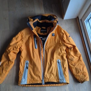 Orange vindjacka från Gneis med huva - Snygg orange vindjacka från Gneis med huva och flera praktiska fickor med dragkedja. Jackan har svarta och grå detaljer, justerbara ärmslut och logga på både ärm och rygg. Perfekt för dig som gillar att vara ute och vill ha en cool och funktionell jacka.