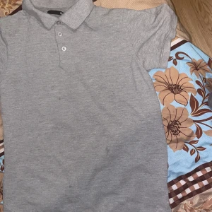 Grå pikétröja från Lager 157 - Grå pikétröja från Lager 157 i storlek M. Tröjan har klassisk krage, tre knappar framtill och korta ärmar. Materialet är mjuk bomull som känns skönt mot huden. Perfekt för dig som gillar enkel och stilren look.