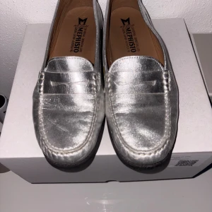 Silvriga loafers från Mephisto - Snygga loafers i metallic silver från Mephisto med klassisk rund tå och platt sula. Skorna är tillverkade i skinn och har en stilren design med dekorativa sömmar och detalj på ovansidan. Perfekta för dig som vill sticka ut med en glansig look. Köpta i New York, originalpris 3500