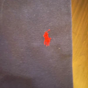 Mörkblå pikétröja från Ralph Lauren - Snygg mörkblå pikétröja från Ralph Lauren med klassisk krage och två knappar framtill. Tröjan har det ikoniska röda Polo-logot broderat på bröstet. Tillverkad i mjuk bomull som är skön mot huden och passar perfekt till jeans eller shorts.