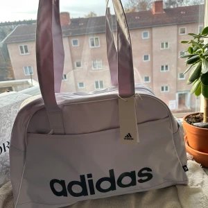 Ljuslila axelväska från Adidas - Snygg ljuslila axelväska från Adidas med stort svart logotryck på framsidan. Väskan har breda axelband och en praktisk ytterficka. Perfekt storlek för att få med allt du behöver och tillverkad i slitstark polyester.