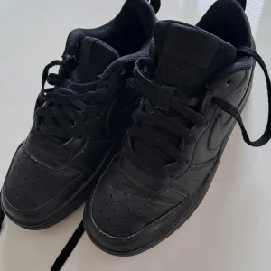 Svarta Nike Air Force 1 sneakers - Klassiska helsvarta Nike Air Force 1 sneakers i skinn med snörning och rund tå. Snygg och tidlös modell med perforerade detaljer på tån och diskret Nike-logga på sidan. Perfekta för dig som gillar en clean och urban stil.