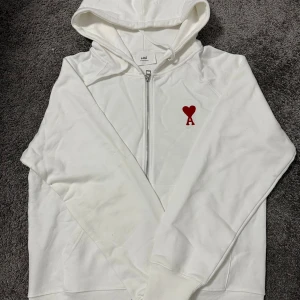 Vit hoodie Ami - Vit hoodie från Ami med dragkedja och huva. På bröstet finns ett broderat rött hjärta med bokstaven A. Tröjan har långa ärmar, ribbade muddar och är tillverkad i mjuk bomull. Snygg och clean design som sticker ut med den röda detaljen.