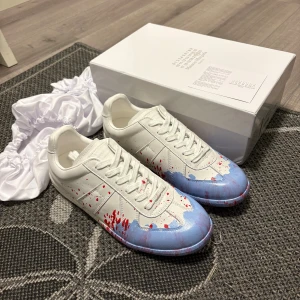 Maison Margiela sneakers blå/röd - Maison Margiela sneakers i vitt skinn med unika blå och röda färgstänk över tån och sidorna. Klassisk låg modell med snörning och rund tå. Kommer med originalkartong och skyddande dustbags. Perfekta för dig som gillar statement-detaljer och exklusiv design.