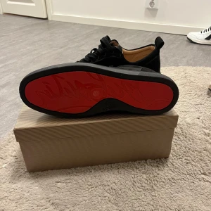  Christian Louboutin - Säljer ett par svarta sneakers från Christian Louboutin i mocka med klassisk röd sula och diskret logga. Skorna har rund tå, snörning och svarta detaljer. Insidan är beige och sulan har Louboutins signatur. Perfekta för dig som gillar lyxiga och stilrena sneakers.