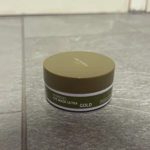 Hydrogel Eye Mask Ultra Gold - Hydrogel Eye Mask Ultra Gold från Rexaline i en rund burk med olivgrönt lock. Innehåller skimrande guldiga ögonmasker och en vit plastspatel för applicering. Perfekt för att pigga upp trötta ögon och ge en fräsch känsla.