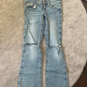 Blå bootcut jeans från Gina Tricot - Snygga ljusblå jeans från Gina Tricot i bootcut-modell med slitna detaljer och hål vid knäna. Klassisk femficksdesign, normal passform och tillverkade i jeansmaterial. Perfekta för dig som gillar en avslappnad och trendig look.
