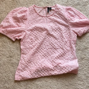 Ljusrosa blus med puffärm från Mango - Superfin ljusrosa blus från Mango med puffiga korta ärmar och rund hals. Blusen har ett strukturerat rutigt mönster i tyget och knapp i nacken. Perfekt för dig som gillar söta detaljer och mjuka färger.