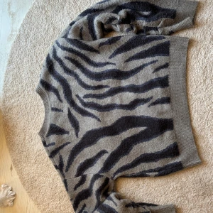 Grå zebramönstrad stickad tröja - Cool stickad tröja från Gina Tricot i grått med svart zebramönster. Rund hals och ribbade muddar vid ärmslut och nederkant. Perfekt för dig som gillar djurmönster och vill ha en mysig, trendig look. Passar dig som vill sticka ut lite extra.