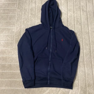 Marinblå hoodie från Polo Ralph Lauren - Snygg marinblå hoodie från Polo Ralph Lauren med dragkedja och huva. Tröjan har snörning vid huvan, två fickor fram och ett litet rött Polo-logo broderat på bröstet. Perfekt för chill dagar och enkel att matcha med jeans eller joggers.