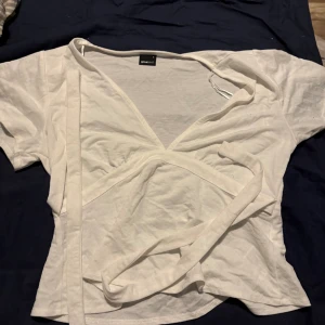 Vit omlottopp från Gina Tricot - Säljer en vit omlottopp från Gina Tricot med korta ärmar och v-ringad hals. Toppen har långa knytband i midjan,men man kan även knyta den i ryggen så det blir en fin rosett, som ger en snygg och justerbar passform. Perfekt för dig som gillar stilrena och enkla plagg med en twist.
