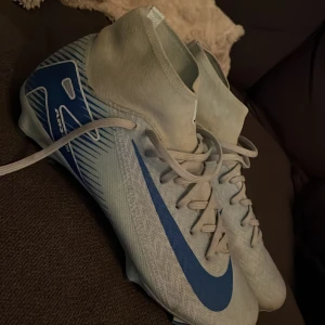Nike Mercurial vita och blå fotbollsskor - Nike Mercurial fotbollsskor i ljusblått med blå detaljer och snörning. Skorna har hög strumpliknande krage, platt sula med dobbar och ett stilrent mönster på hälen. Perfekta för dig som vill ha grym kontroll och snabbhet på planen. Skon har syntetiskt underlag och är multiground. Har inte alls användt dessa länge då jag slutade i sporten. Men dessa är mina absoluta favorit fotbollsskor jag haft, rekommenderar starkt. Nypris 1300kr