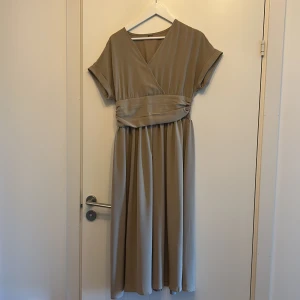 Beige omlottklänning i polyester - Snygg beige långklänning med omlottliv och bred markerad midja. Klänningen har korta ärmar, v-ringning och är tillverkad i 100% polyester. Perfekt för dig som gillar en stilren och flowy look.