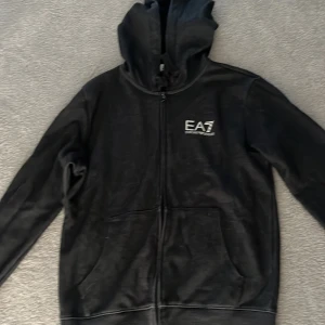 Svart EA7 Emporio Armani hoodie - Svart hoodie från EA7 Emporio Armani med dragkedja framtill och vit logga på bröstet. Tröjan har huva, långa ärmar och två fickor framtill. Snygg och stilren design med diskreta vita detaljer även på ryggen.