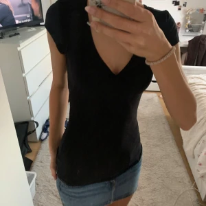 Svart v-ringad topp med kort ärm - Säljer en svart topp med v-ringning och korta ärmar. Toppen har en lätt twistad detalj vid bysten och sitter figurnära. Perfekt att styla med jeans eller kjol för en clean look. Ett hål vid armhålan 
