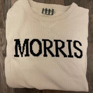 Vit stickad tröja från Morris - Snygg vit stickad tröja från Morris med svart logotyp broderad över bröstet. Tröjan har rund hals, ribbade muddar och lång ärm. Perfekt för dig som gillar klassisk stil med tydlig branding.