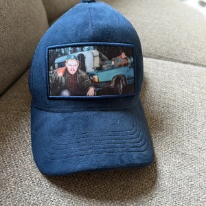 Blå meshkeps från Wristwear - Säljer en blå keps från Wristwear med mesh på baksidan och justerbar snapback. Framsidan har en unik patch med bildmotiv av två personer och en blå bil. Kepsen har mjuk mockaliknande yta framtill och cool streetstil.