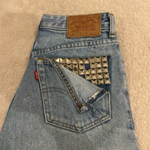 Levi's 501 ljusblå jeansshorts med nitar - Ljusblå Levi's 501 jeansshorts med hög midja och råa, fransiga benslut. Shortsen har silvriga pyramidnitar på fram- och bakficka för en edgy vibe. Klassisk femficksmodell i slitstark bomullsdenim. Perfekta för dig som vill sticka ut lite extra.