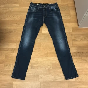 Replay Anbass Hyperflex jeans blå 32/32 - Snygga Replay Anbass Hyperflex jeans i mörkblå tvätt med diskreta slitningar och klassiska femficksdetaljer. Stretchigt material för extra komfort och smal passform. Märkespatch i läder baktill och tydliga kontrastsömmar. Perfekta för en trendig och avslappnad look.
