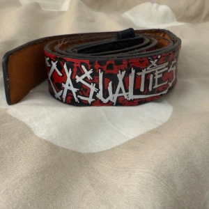The Casualties bälte med dödskallar - Säljer ett svart och rött bälte från The Casualties med coolt tryck av vita bokstäver och röda dödskallar. Bältet är gjort i skinn och har en punkig vibe som sticker ut. Perfekt accessoar för dig som gillar alternativ stil och vill ha något unikt i garderoben.