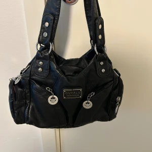 Svart axelväska från Ouyalu - Cool svart axelväska i syntetmaterial med breda handtag, silvriga nitar och dragkedjor. Väskan har flera fack med snygga metallhängen och detaljer framtill. Perfekt för dig som gillar en edgy och praktisk look. Rymmer allt du behöver till vardagen.