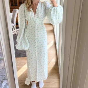 Vit långklänning med blommigt mönster - Superfin vit långklänning med ljusblått blommigt mönster och v-ringning. Klänningen har puffiga långärmar, knappar längs hela framsidan och markerad midja. 