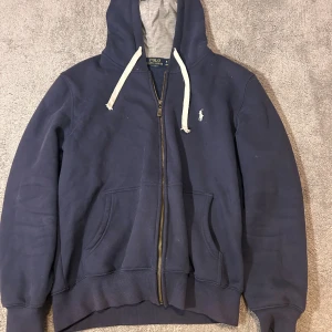Marinblå hoodie från Polo Ralph Lauren - Marinblå hoodie från Polo Ralph Lauren med vit dragsko och dragkedja framtill. Klassisk logga broderad på bröstet och två stora fickor. Mjuk insida och huva med grått foder. Perfekt för chill dagar eller när du vill ha en clean streetstyle.