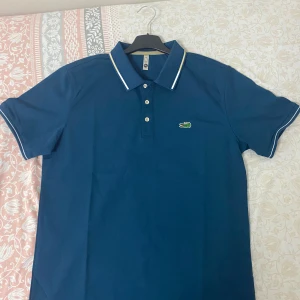 Blå pikétröja från Lacoste - Det står xl men är MSnygg blå pikétröja från Lacoste med klassisk krage, tre knappar och den ikoniska krokodilloggan på bröstet. Tröjan har vita detaljer på krage och ärmslut och är gjord i mjuk bomull. Perfekt för en stilren och sportig look.