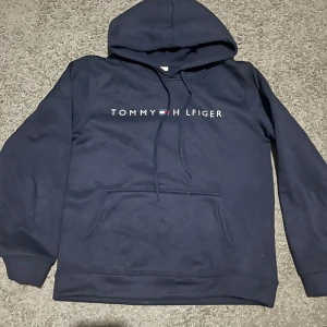 Marinblå hoodie från Tommy Hilfiger - Snygg marinblå hoodie från Tommy Hilfiger med klassisk logga framtill. Tröjan har huva med snörning och en stor magficka. Perfekt för dig som gillar stilrena och bekväma plagg med streetkänsla.