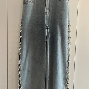 Ljusblå jeans med strassdetaljer - Unika ljusblå jeans med raka ben och bred passform. Sidorna är öppna och dekorerade med glittrande strasskedjor som ger en riktigt cool och trendig look. Perfekta för dig som vill sticka ut och älskar detaljer som syns.