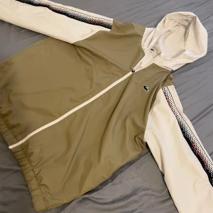 Beige och vit vindjacka från Lacoste Sport - Snygg vindjacka från Lacoste Sport i beige och vitt med huva och hel dragkedja. Jackan har vita ärmar med svart och röd grafisk detalj längs sidorna, samt elastisk nederkant. Klassisk krokodillogga på bröstet och meshfoder för extra komfort.