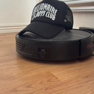 Svart Billionaire Boys Club keps - Snygg svart truckerkeps från Billionaire Boys Club med vit logga framtill. Kepsen har böjd skärm och mesh på sidorna och bak för extra ventilation. Perfekt accessoar för en streetwear-look.