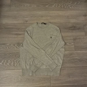 Grå stickad tröja från Polo Ralph Lauren - Snygg grå stickad tröja från Polo Ralph Lauren med klassisk rund halsringning och broderad svart logga på bröstet. Tröjan har långa ärmar och ribbade muddar för en clean look. Perfekt för lager-på-lager under kyliga dagar. Tröjan sitter litet, skyller rekommendera M i storlek om den ska passa. Det är en andel kashmir i tröjan. Det finn inget täncken på användning. Skick 9/10.