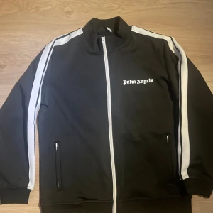 Palm Angels svart track jacket - Svart track jacket från Palm Angels med vita ränder längs ärmarna och vit dragkedja. Jackan har hög krage, två fickor med dragkedja och Palm Angels-logga tryckt på bröstet. Perfekt för en sportig och trendig streetwear-look.