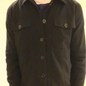 Svart overshirt från Dressmann S - Tja, Säljer nu denna svarta overshirtern från Dressmann med klassisk krage och fyra stora knappar framtill. Perfekt för lager-på-lager och enkel att matcha med det mesta. Säljer den för den ej passar mig längre, Passar perfekt på dig runt 170-175 ungefär. Inga synliga deffekter, Bra skick!
