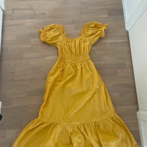 Gul långklänning med puffärmar - Superfin gul långklänning med puffiga kortärmar och broderade detaljer. Klänningen har smock i midjan och volang nedtill för extra flow. Perfekt för dig som vill sticka ut med färg och romantisk stil. Materialet känns lätt och luftigt, idealiskt för varma dagar.