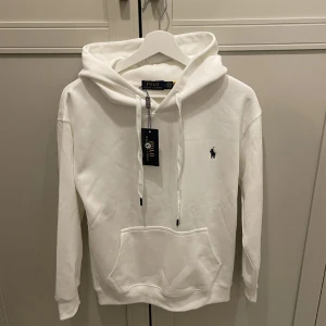 Vit hoodie från Polo Ralph Lauren - Helt ny Vit hoodie från Polo Ralph Lauren med klassisk huva och snörning. Tröjan har en stor magficka och den ikoniska Polo-loggan broderad på bröstet. Perfekt för chill dagar och enkel att matcha med jeans eller joggers. 