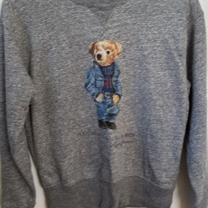 Grå sweatshirt Polo Ralph Lauren XS - Grå sweatshirt från Polo Ralph Lauren med ikonisk broderad nalle på bröstet. Klassisk rund halsringning och långa ärmar. Mjuk och skön bomullsblandning, perfekt för en chill och avslappnad stil.