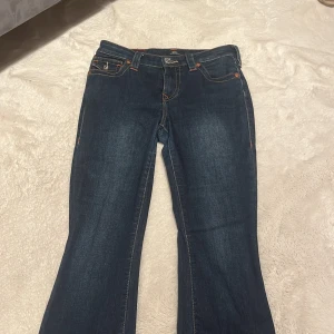 True Religion Becca Bootcut Jeans - Mörkblå jeans från True Religion, modell Becca Mid Rise Bootcut. Snygga kontrastsömmar, klassiska fickor med lock och silverfärgade knappar. Bootcut-modell med medelhög midja och ikonisk True Religion-logga bak.