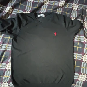 Svart tröja från Ami Paris med rött hjärta - Svart tröja från Ami Paris med klassisk rund hals och ribbade muddar. På bröstet finns det ikoniska broderade röda hjärtat med bokstaven A. Tröjan är i mjuk bomull och har en clean, stilren look som passar perfekt till allt 