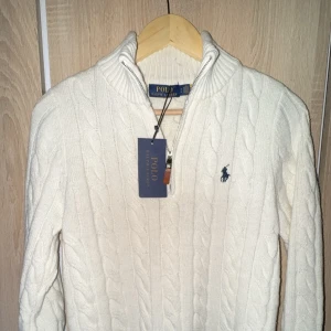 Vit kabelstickad tröja Polo Ralph Lauren - Kabelstickad vit tröja från Polo Ralph Lauren med halv dragkedja och hög krage. Klassisk design med broderad logga på bröstet. Tillverkad i mjukt stickat material, perfekt för kyligare dagar.