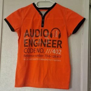 Orange t-shirt från Skill med tryck - Orange t-shirt från Skill med svart tryck 'Audio Engineer' och detaljer som svarta muddar och knappar vid halsen. T-shirten har korta ärmar och är gjord i polyester. Perfekt för dig som vill sticka ut med färg. Hela t-shirten är nopprig men ser varken fläckar eller hål.