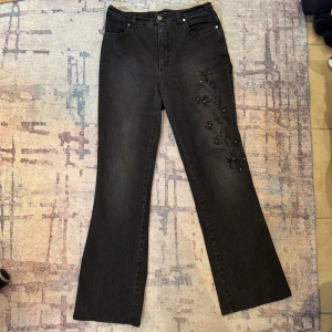 Svarta jeans med broderade blommor - Snygga svart/gråa jeans med broderade blommor samt små pärlor på ena benet. I mycket fint skick! Midjemått - 37cm