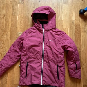 Mörkrosa vindjacka från Everest - Snygg mörkrosa vindjacka från Everest med huva och hel dragkedja framtill. Jackan har två fickor med dragkedja och svart innerfoder. Materialet är nylon och polyester, perfekt för blåsiga dagar. Enkel och sportig design med logga på bröstet.