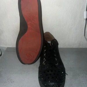 Svarta sneakers med nitar från Louboutin - Säljer ett par svarta sneakers från Louboutin med ikonisk röd sula. Skorna har glittrande ovandel och är täckta med svarta nitar på tå och häl för en edgy look. Klassisk snörning och rund tå. Perfekta för dig som vill sticka ut med en lyxig streetstyle.