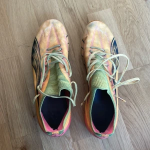 Färgglada Puma fotbollsskor med snörning - Säljer ett par riktigt coola fotbollsskor från Puma i flera färger – orange, rosa och svart. Skorna har snörning, mönstrad ovandel och rosa dobbar undertill. Perfekta för dig som vill sticka ut på planen! Materialet är syntet och skon har en smal passform. Skorna är tillverkade för spel på både naturgräs och konstgräs.